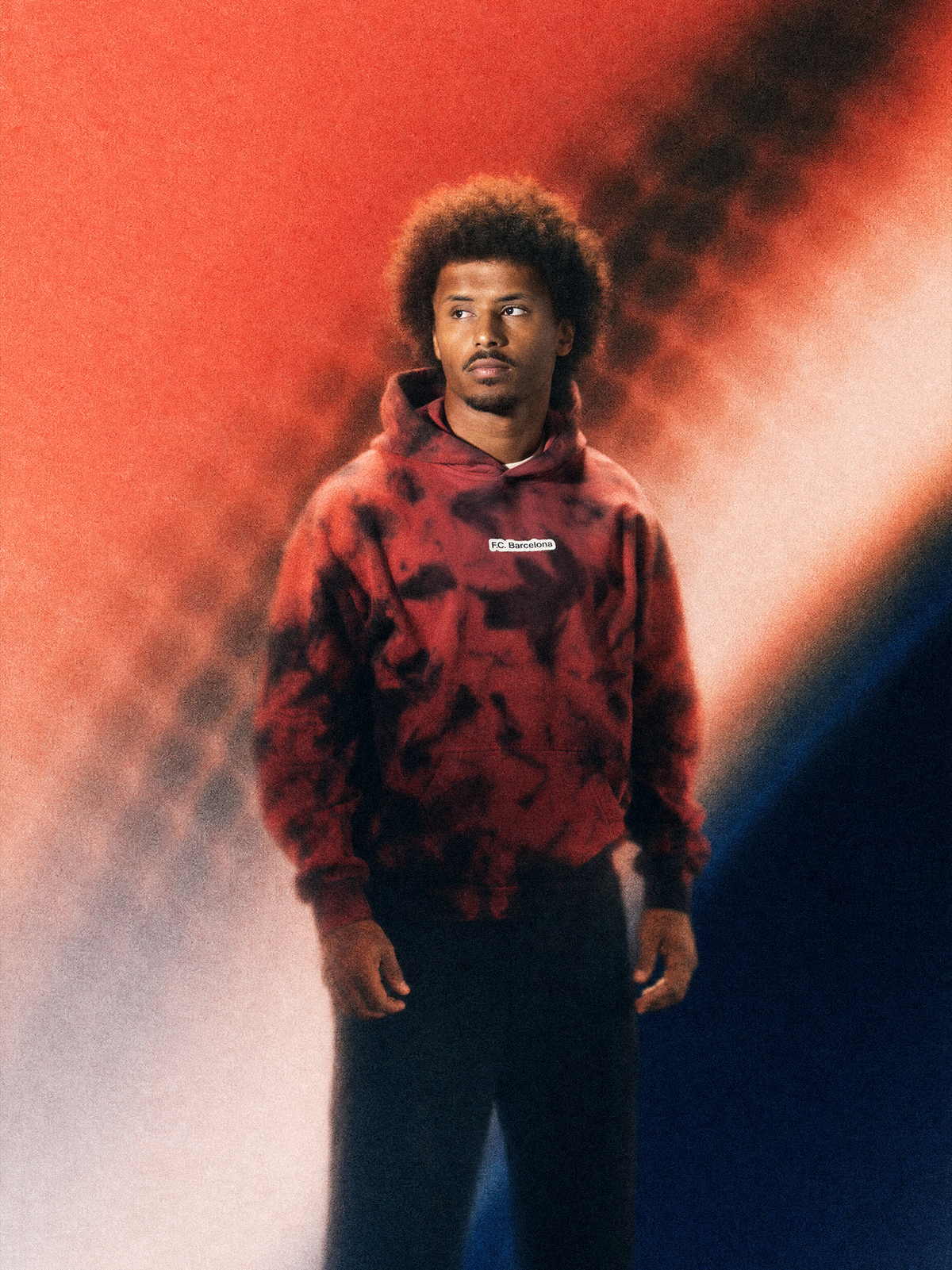 Modelo con sudadera FC Barcelona tie-dye roja y negra, con pequeño logo en el pecho y pantalón negro.