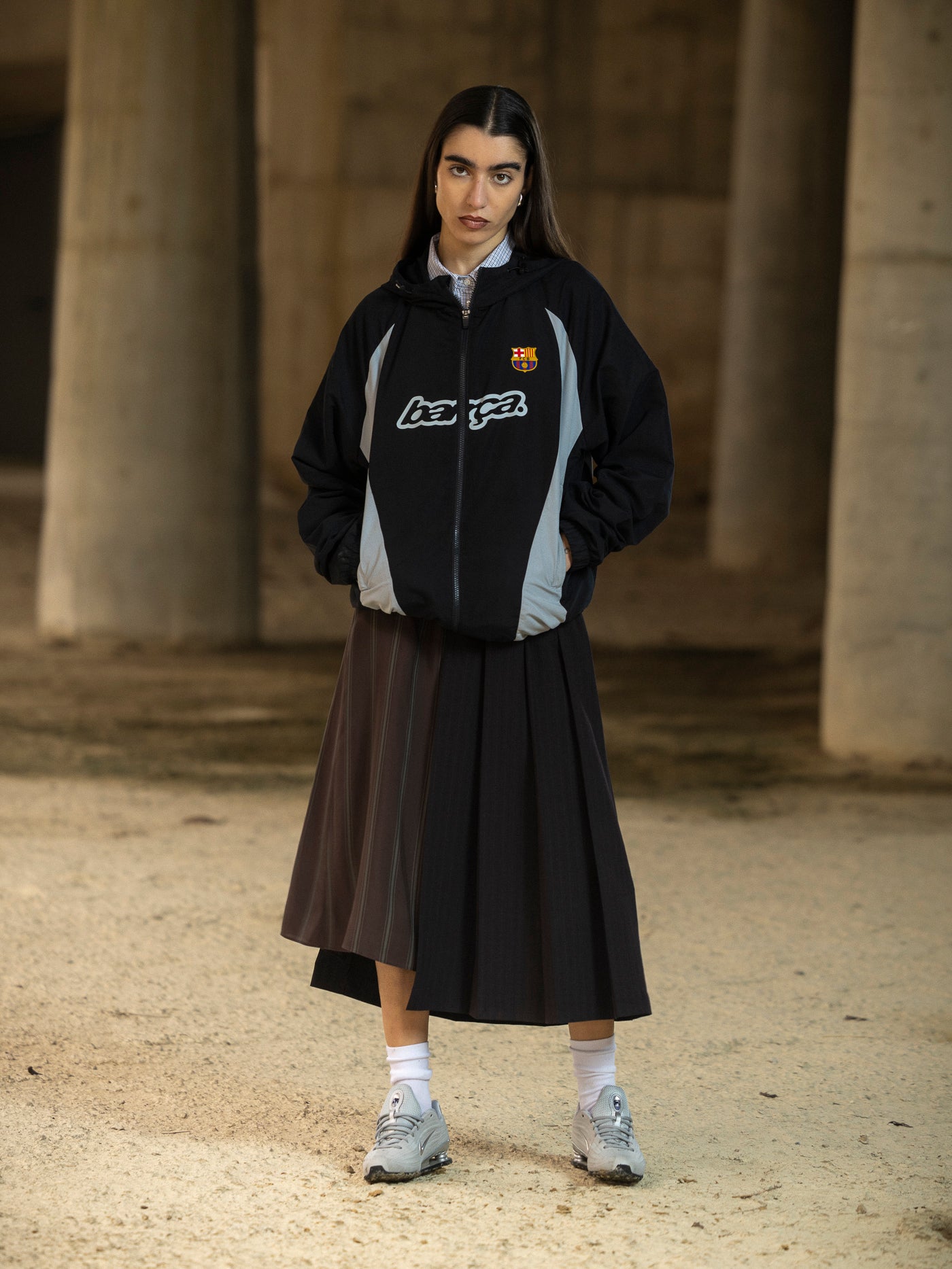 Person wearing a black jacket with a logo and a brown skirt standing in an outdoor setting.