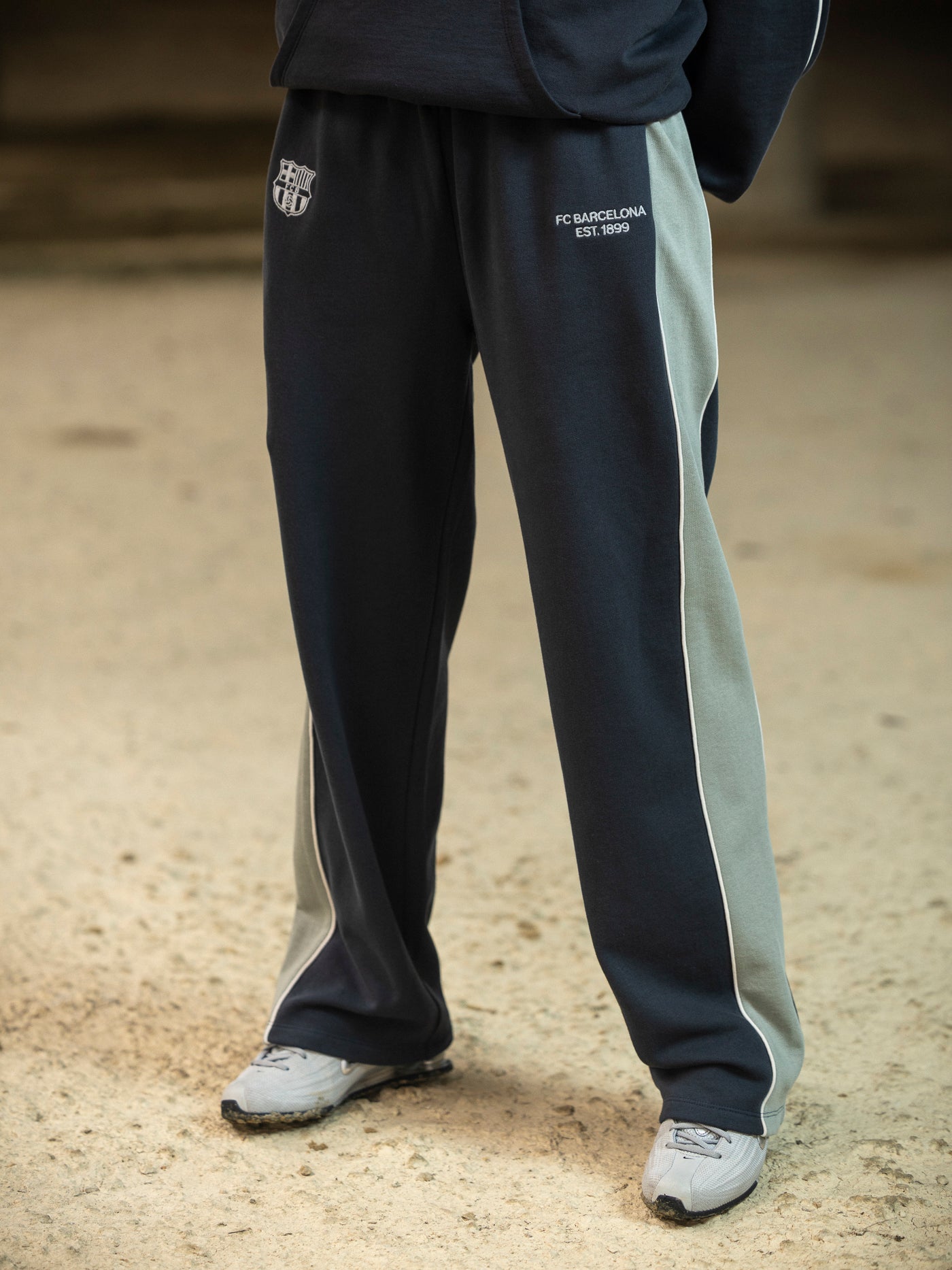 Person wearing dark blue and light gray tracksuit pants with visible branding on a sandy ground.