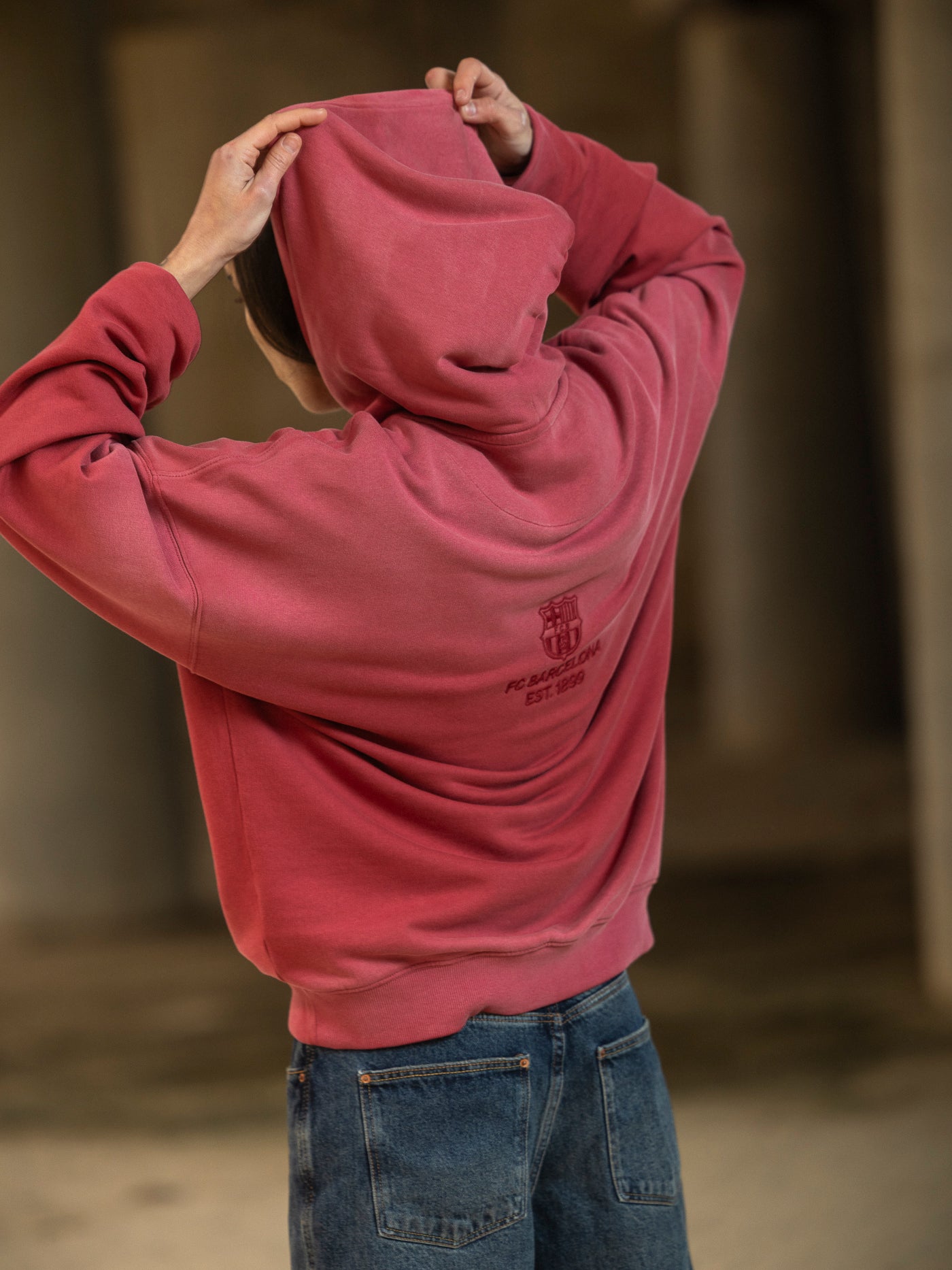 Person wearing a red hoodie with a logo on the back, standing in a blurred indoor setting.