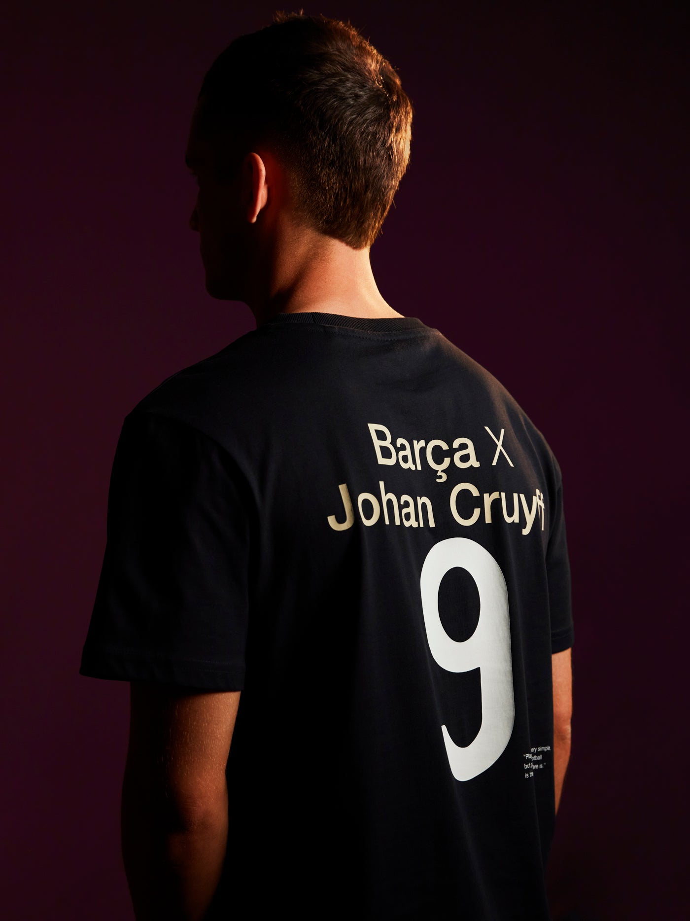 Person wearing a black T-shirt featuring 'Barça X Johan Cruyff' and the number 9 printed on the back, in a dimly lit setting with a purple background.