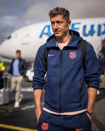 Robert Lewandowski wearing a navy blue FC Barcelona Nike zip-up hoodie with coral crest and swoosh, carrying a black backpack.