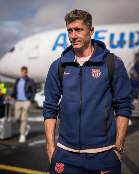 Robert Lewandowski wearing a navy blue FC Barcelona Nike zip-up hoodie with coral crest and swoosh, carrying a black backpack.
