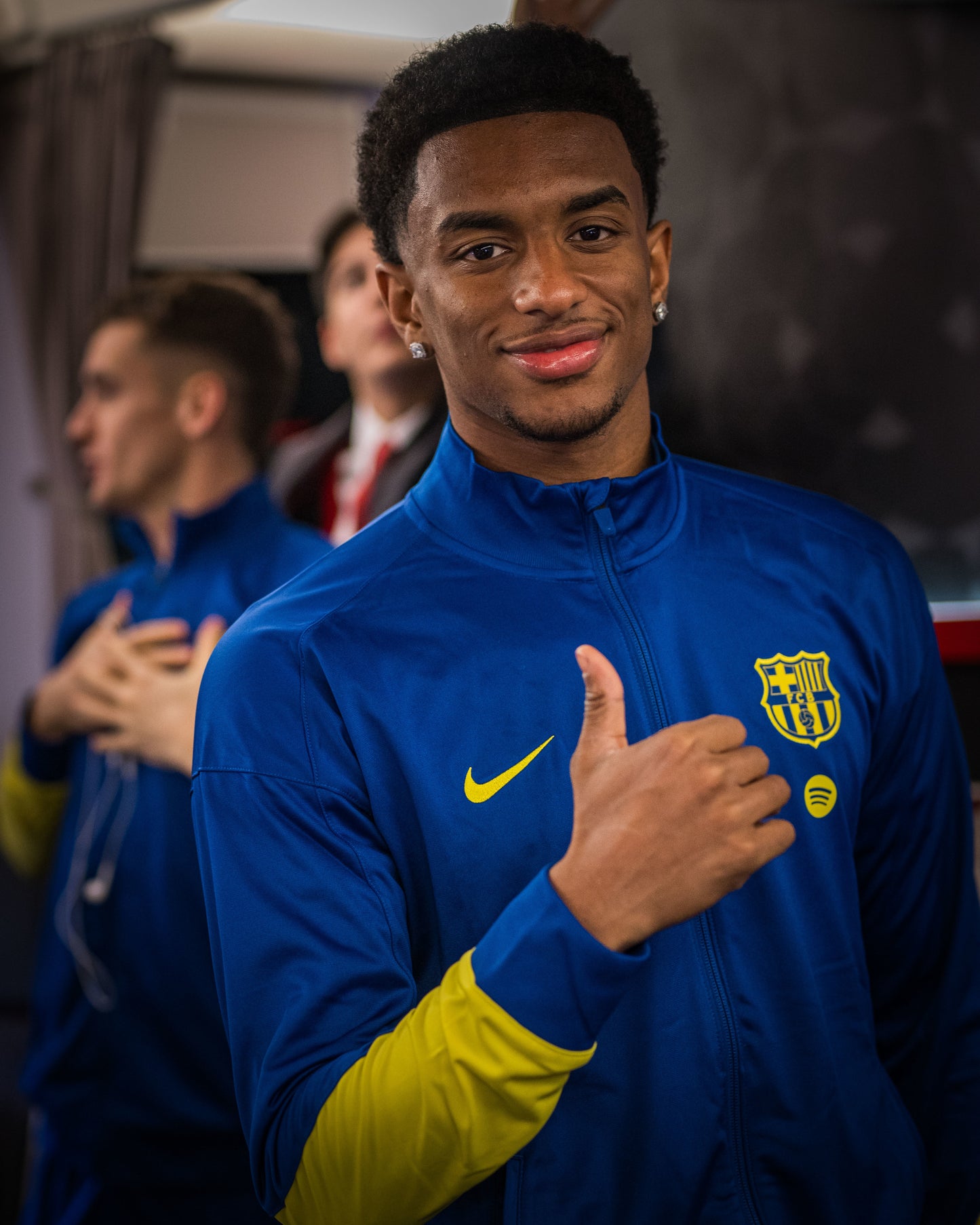 Person wearing a blue sports jacket with a logo, standing in a room with other people.