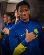 Person wearing a blue sports jacket with a logo, standing in a room with other people.