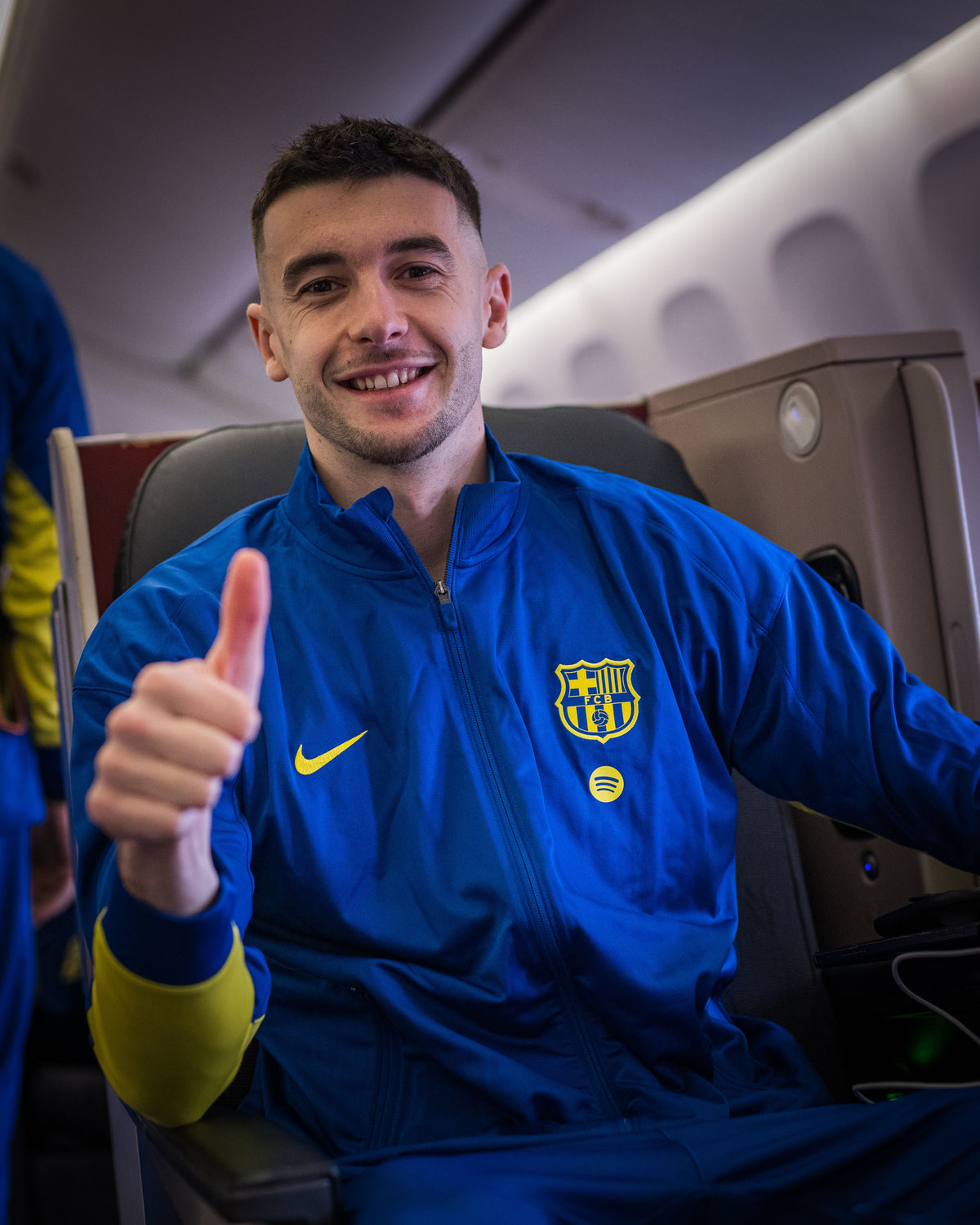 Person wearing a blue sports jacket with a logo, sitting in an airplane.