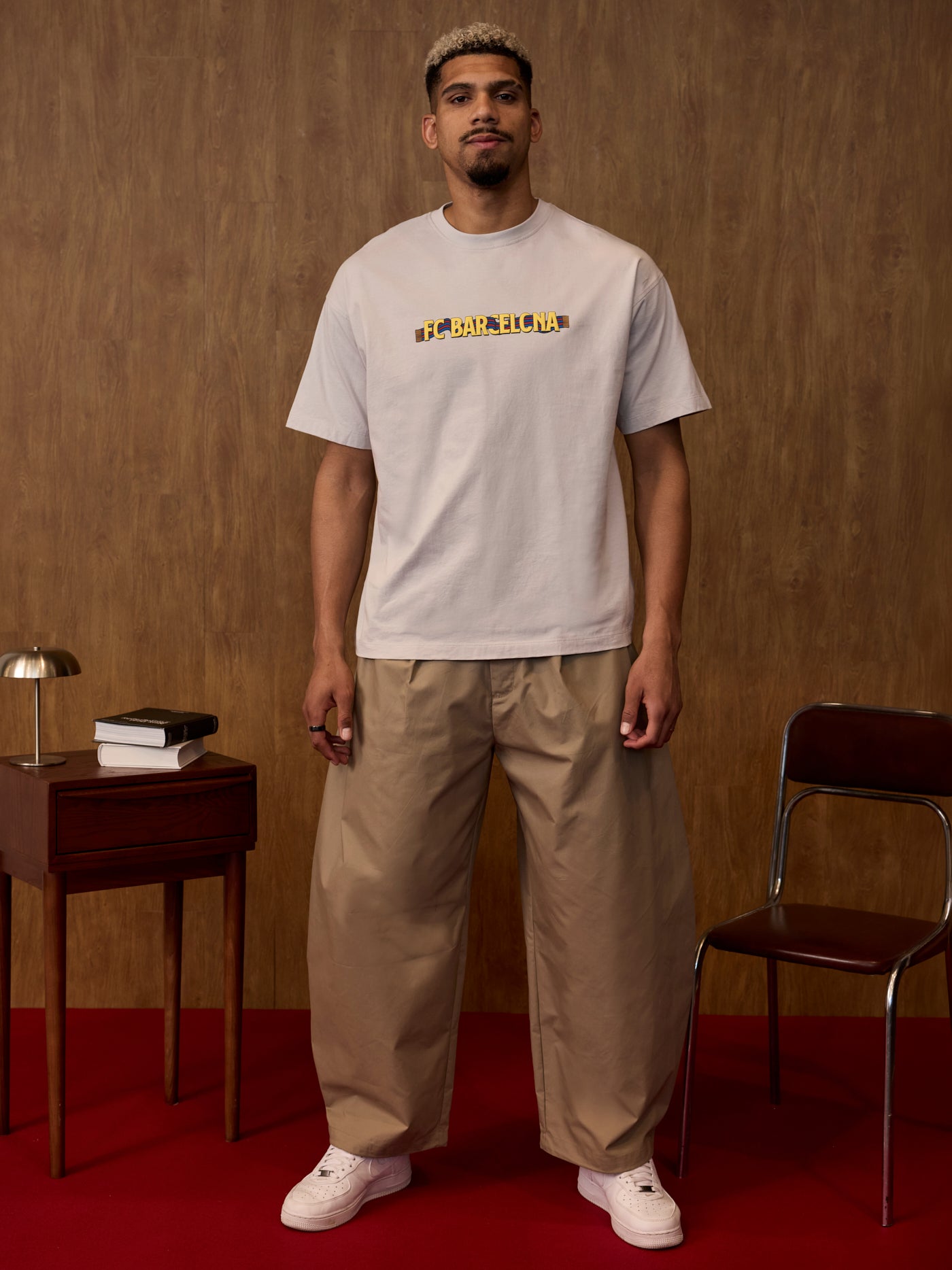 Man wearing a light gray t-shirt with text and beige pants standing in a room with a wooden wall and red floor.