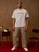 Man wearing a light gray t-shirt with text and beige pants standing in a room with a wooden wall and red floor.