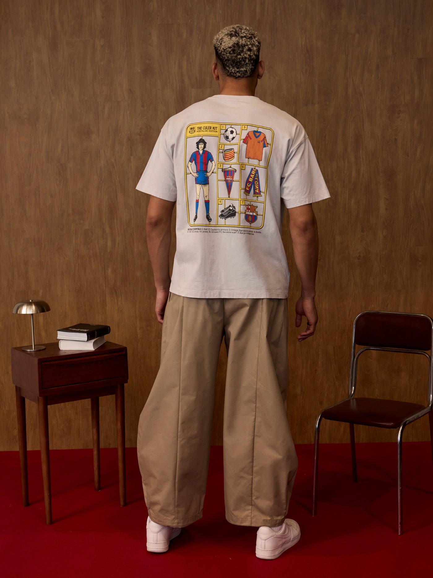 Person wearing a white t-shirt with a colorful graphic on the back, standing in a room with a wooden wall and red floor.