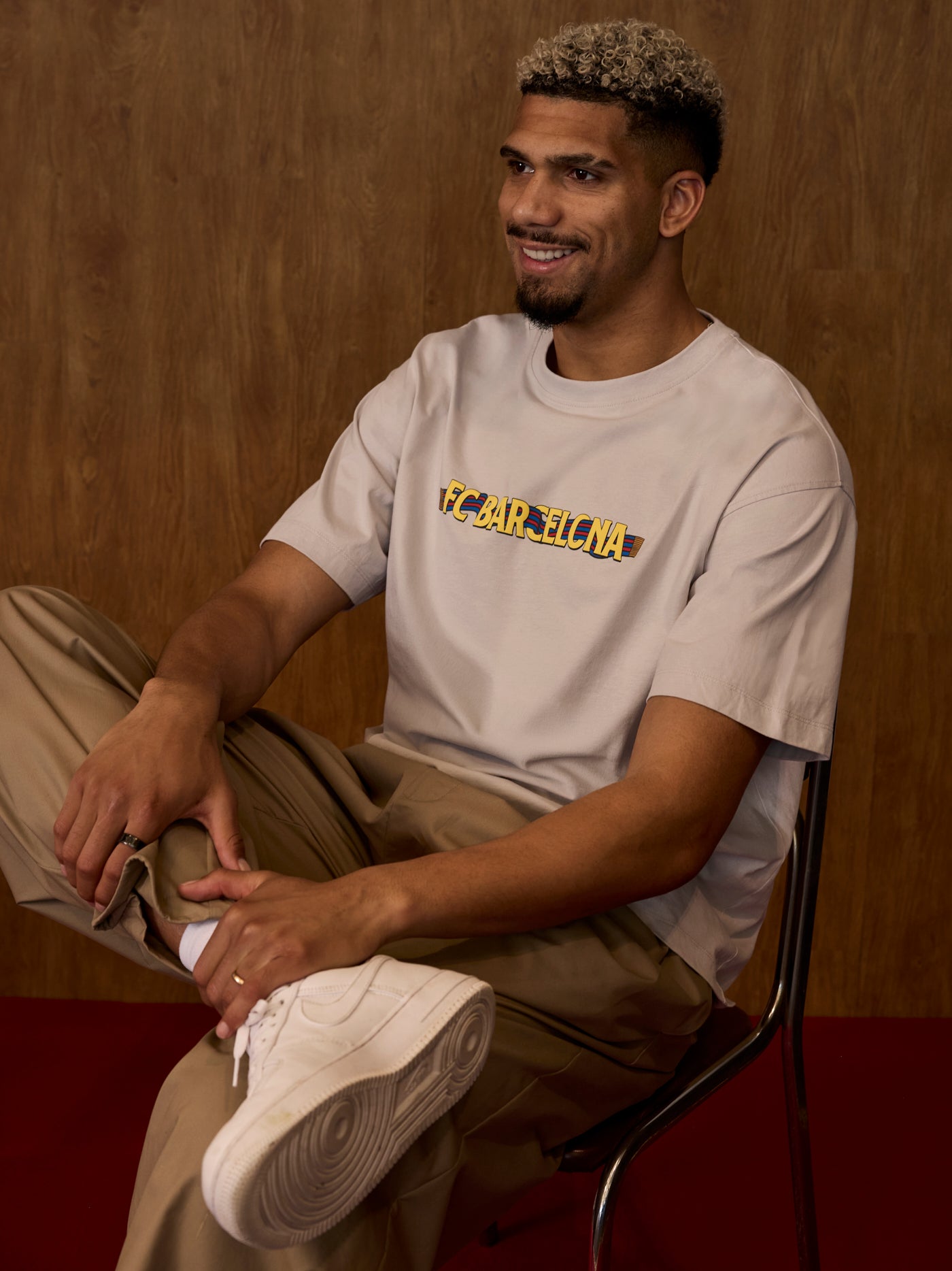 Man sitting on a chair wearing a beige t-shirt with text and light-colored pants.