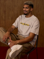 Man sitting on a chair wearing a beige t-shirt with text and light-colored pants.