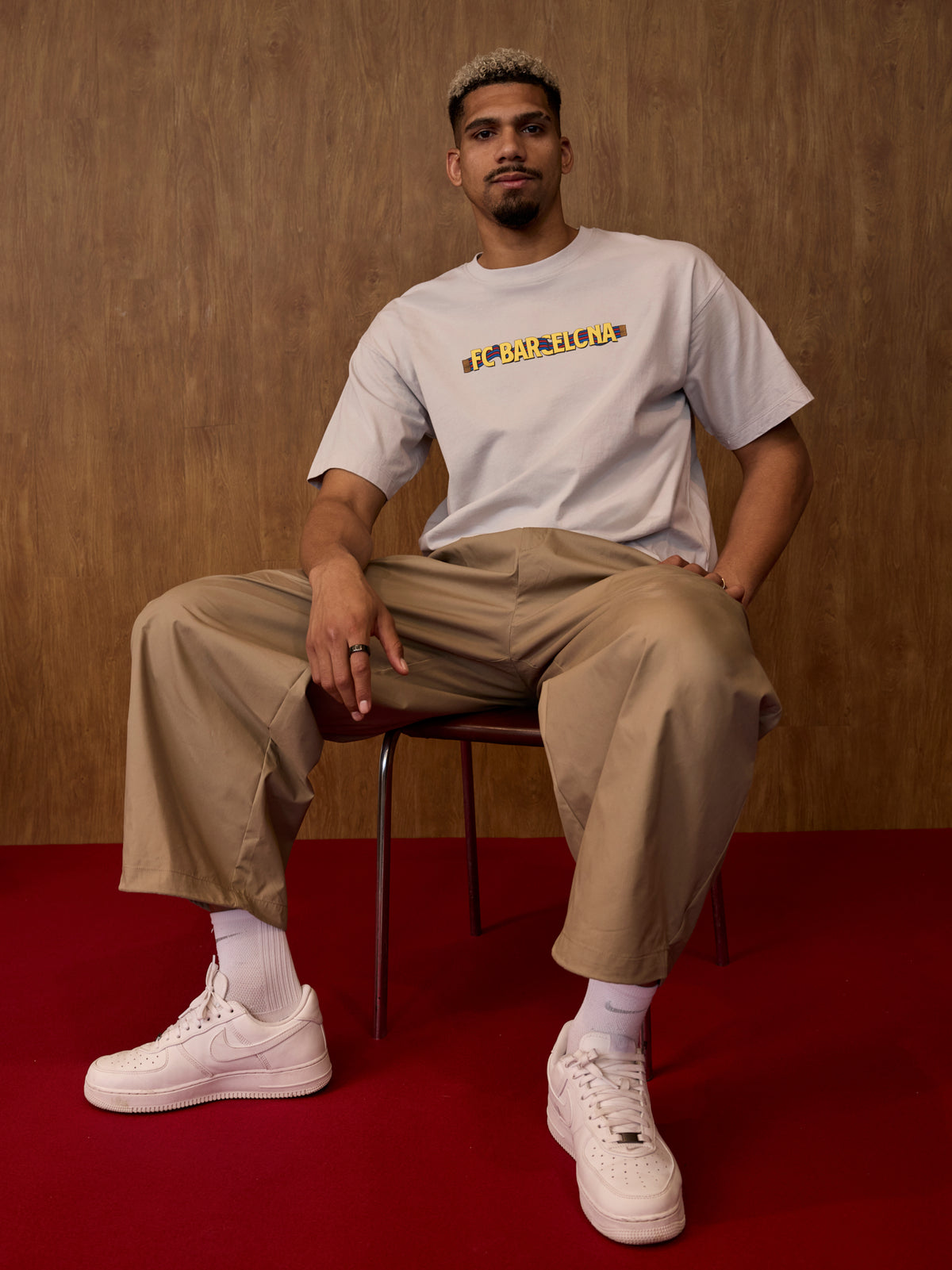 Man sitting on a chair wearing a white t-shirt with text, beige pants, and white sneakers against a brown background.