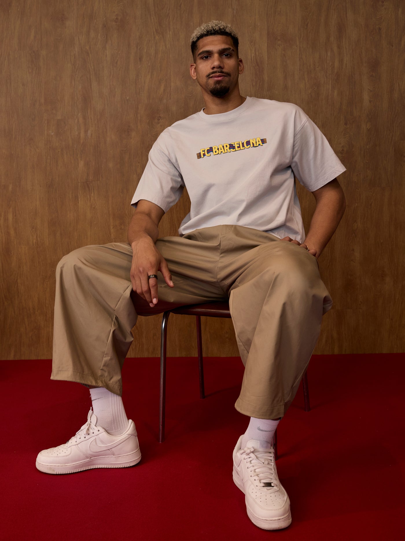 Man sitting on a chair wearing a white t-shirt with text, beige pants, and white sneakers against a brown background.