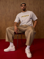 Man sitting on a chair wearing a white t-shirt with text, beige pants, and white sneakers against a brown background.
