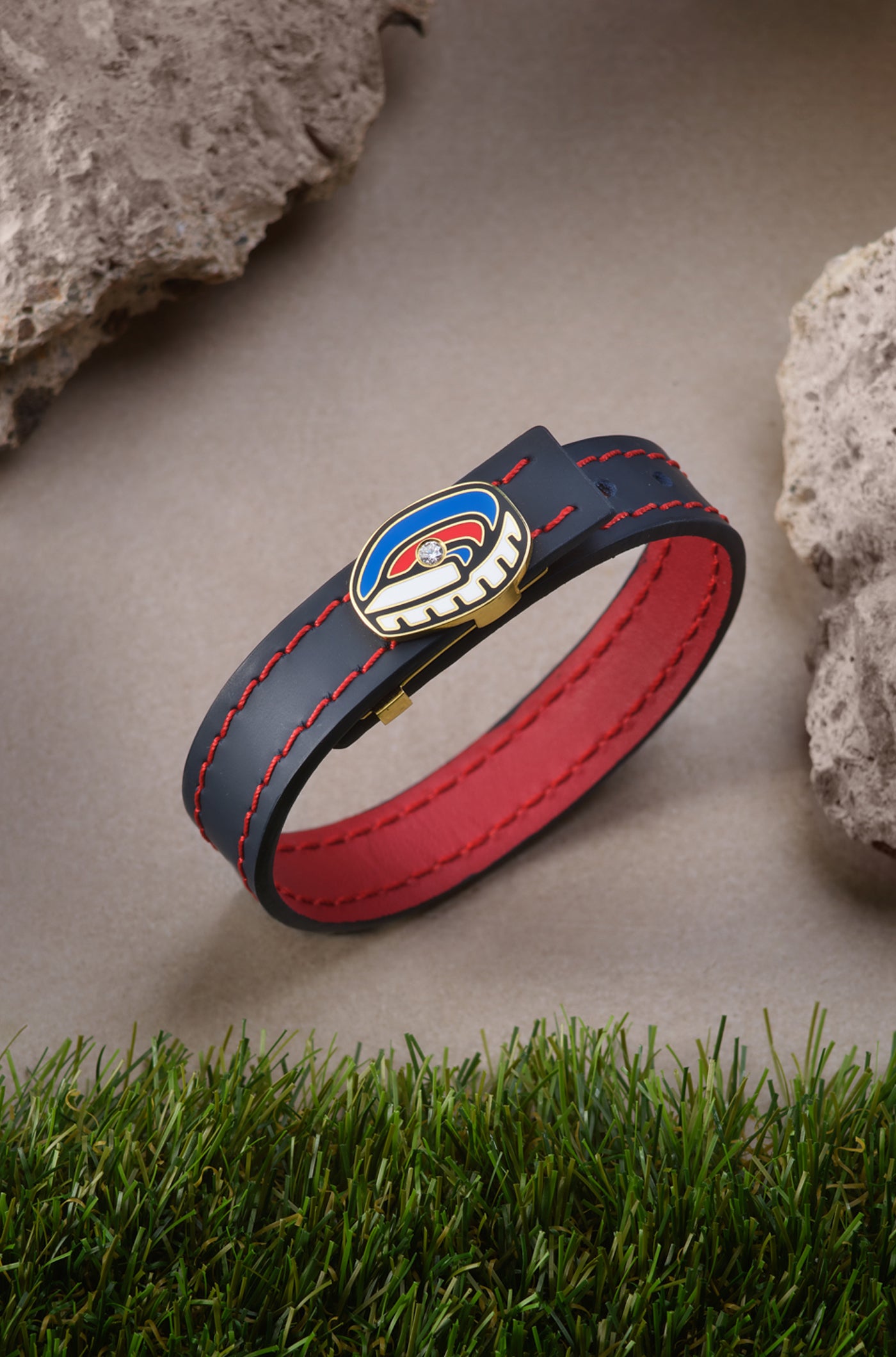 Black leather bracelet with red stitching and red inner lining, featuring a gold buckle shaped like the Spotify Camp Nou stadium in blue, red, and white, with a sparkling gemstone in the center.


