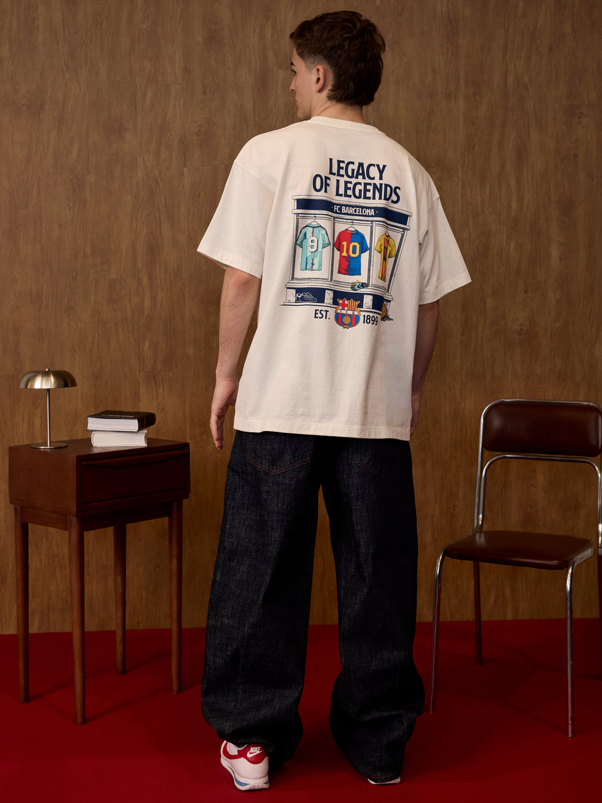 Person wearing a white t-shirt with a graphic design, standing in a room with a wooden wall and red floor.