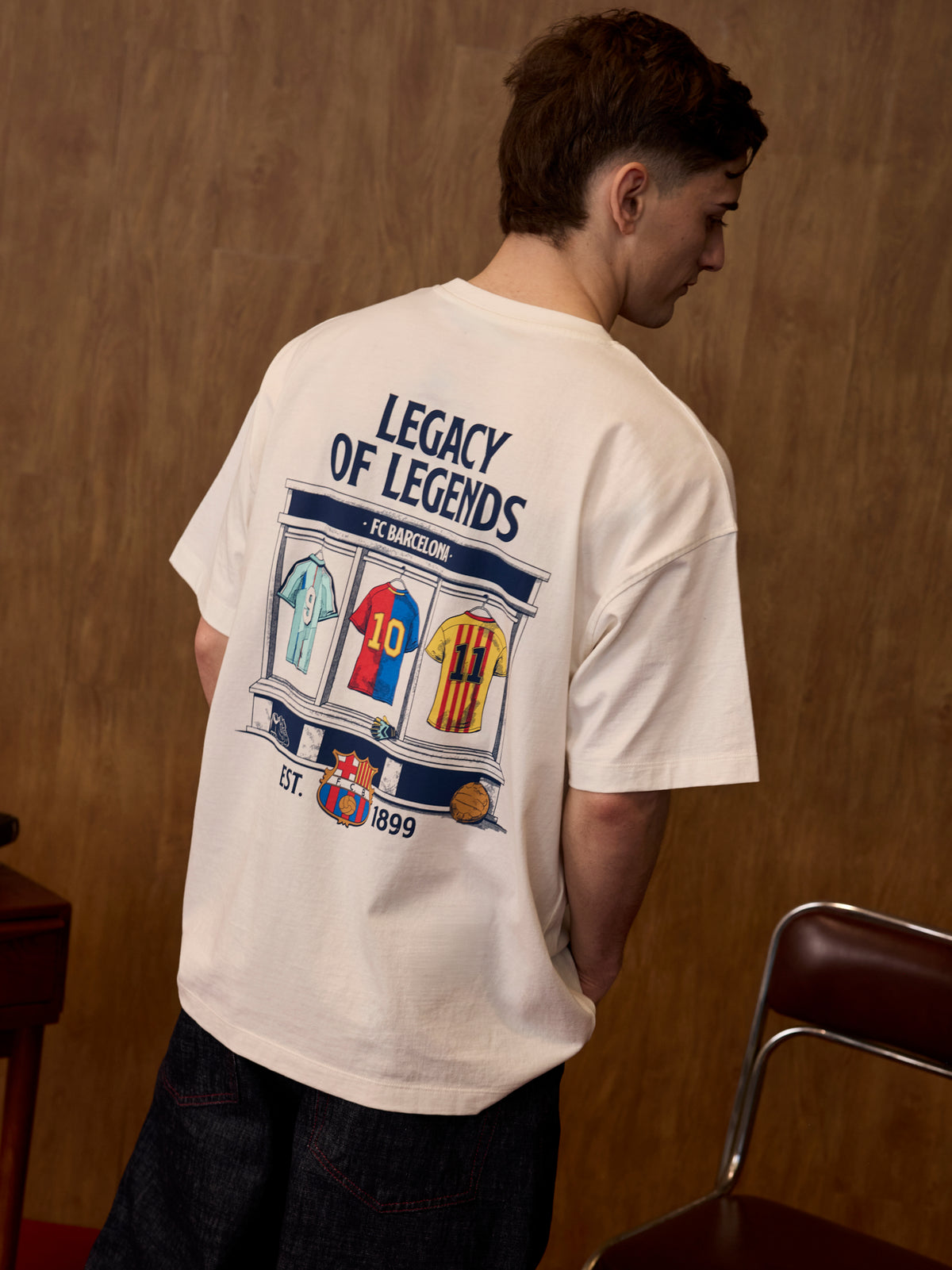 Person wearing a white t-shirt with 'Legacy of Legends' design on the back, standing against a wooden wall.