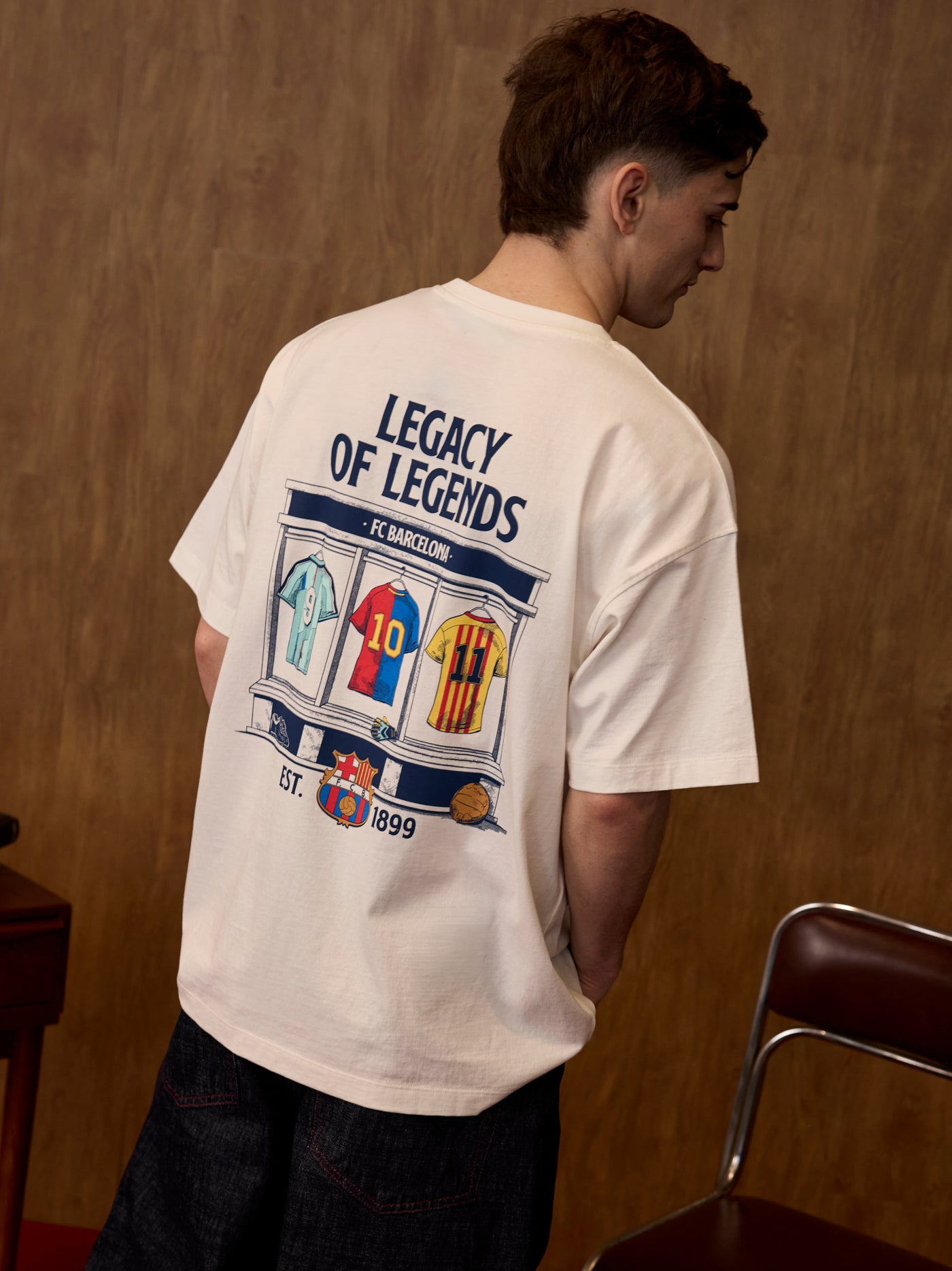 Person wearing a white t-shirt with 'Legacy of Legends' design on the back, standing against a wooden wall.