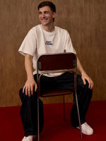 Person sitting on a chair wearing a white t-shirt with text, against a brown wall.