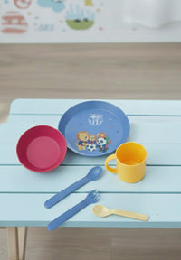 Children's tableware set with plate, bowl, cup, and utensils on a light blue wooden surface.
