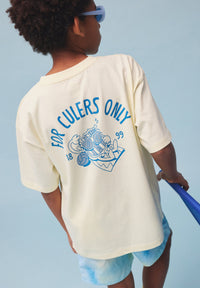 Child wearing a cream t-shirt with blue text and graphics, standing against a light blue background.