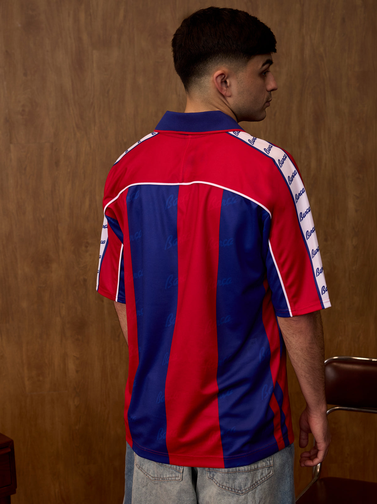 Vista posterior d’una samarreta de FC Barcelona sobre un model, amb franges verticals vermelles i blaves i cinta blanca a les espatlles amb text Barça repetit