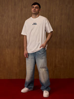 Man wearing a white t-shirt with text and blue jeans against a brown background