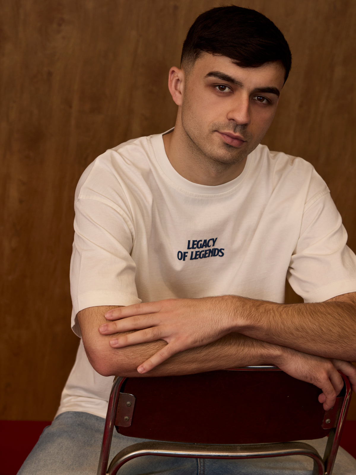 Man wearing a white t-shirt with 'Legacy of Legends' text, sitting on a chair against a brown background