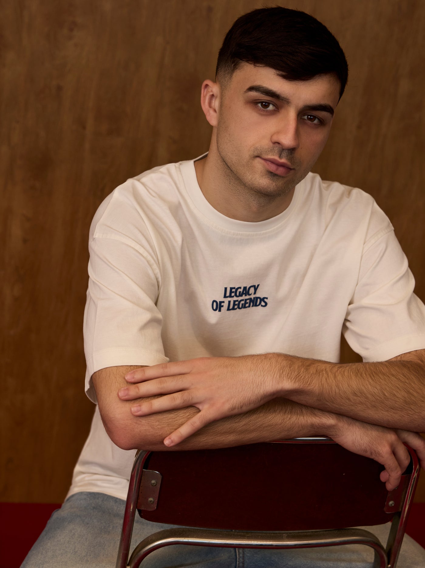Man wearing a white t-shirt with 'Legacy of Legends' text, sitting on a chair against a brown background