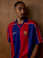 Vista frontal d’una samarreta del FC Barcelona sobre un model, amb franges verticals vermelles i blaves, coll blau i l’escut del club al pit.