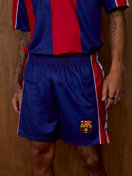 Vue de face d'un équipement de FC Barcelona porté par un modèle, avec un maillot rayé bleu et rouge et un short bleu assorti, avec l'écusson du club.