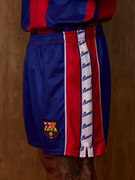 Vue latérale du short FC Barcelona en bleu et rouge, avec une bande verticale script Barça et l’écusson du club sur la jambe.