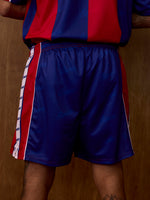 Vue de face du short FC Barcelona bleu avec empiècements latéraux rouges et bordures blanches, porté avec le maillot rayé assorti.