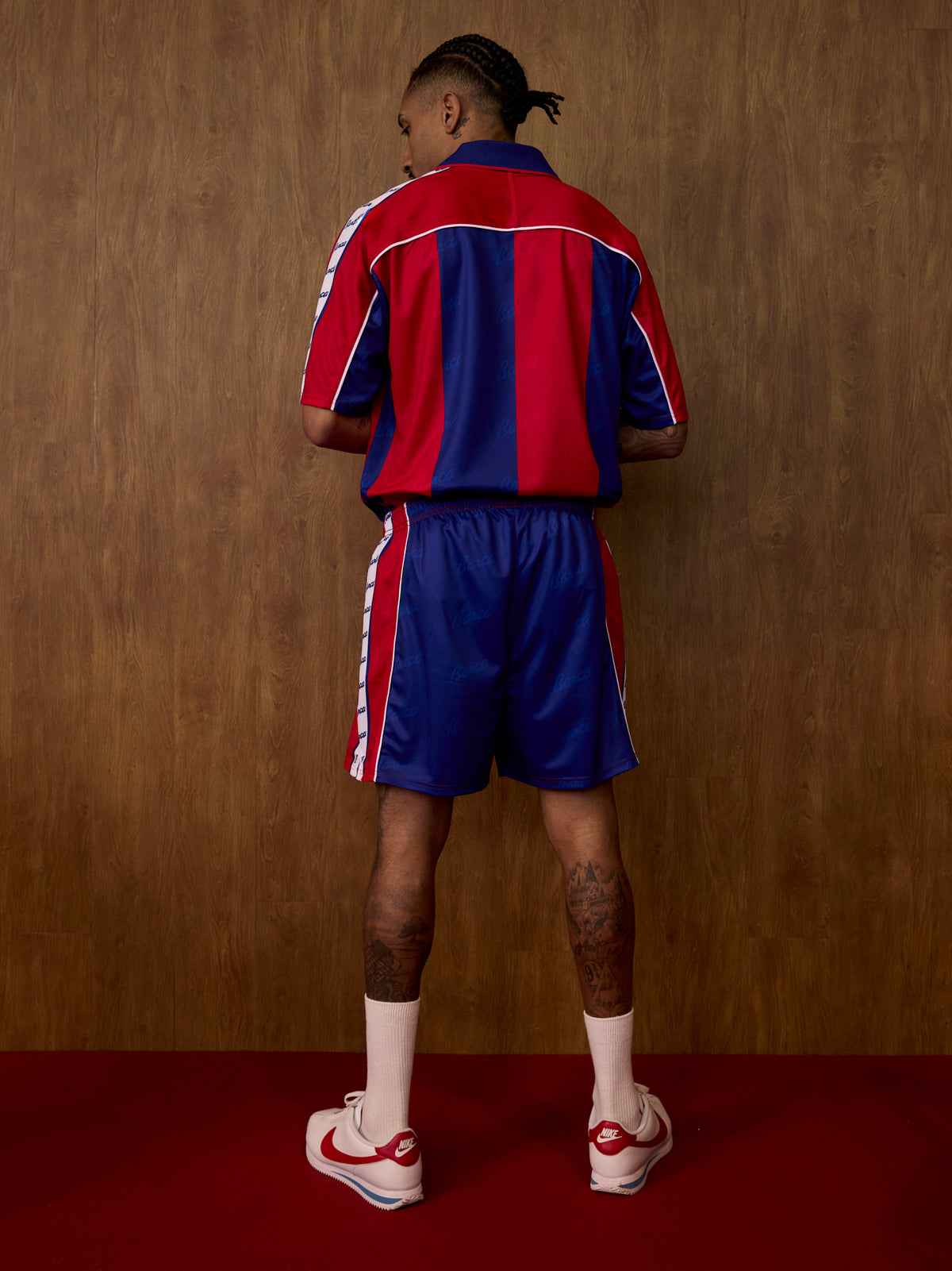 Vista posterior d'un equipament de FC Barcelona sobre un model, amb samarreta de ratlles vermelles i blaves, pantalons curts a joc, rivets blancs i mitjons blancs.