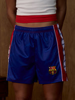 Vista frontal d'uns pantalons curts blaus de FC Barcelona amb franges laterals vermelles i blanques i l'escut del Barça a la cama dreta.