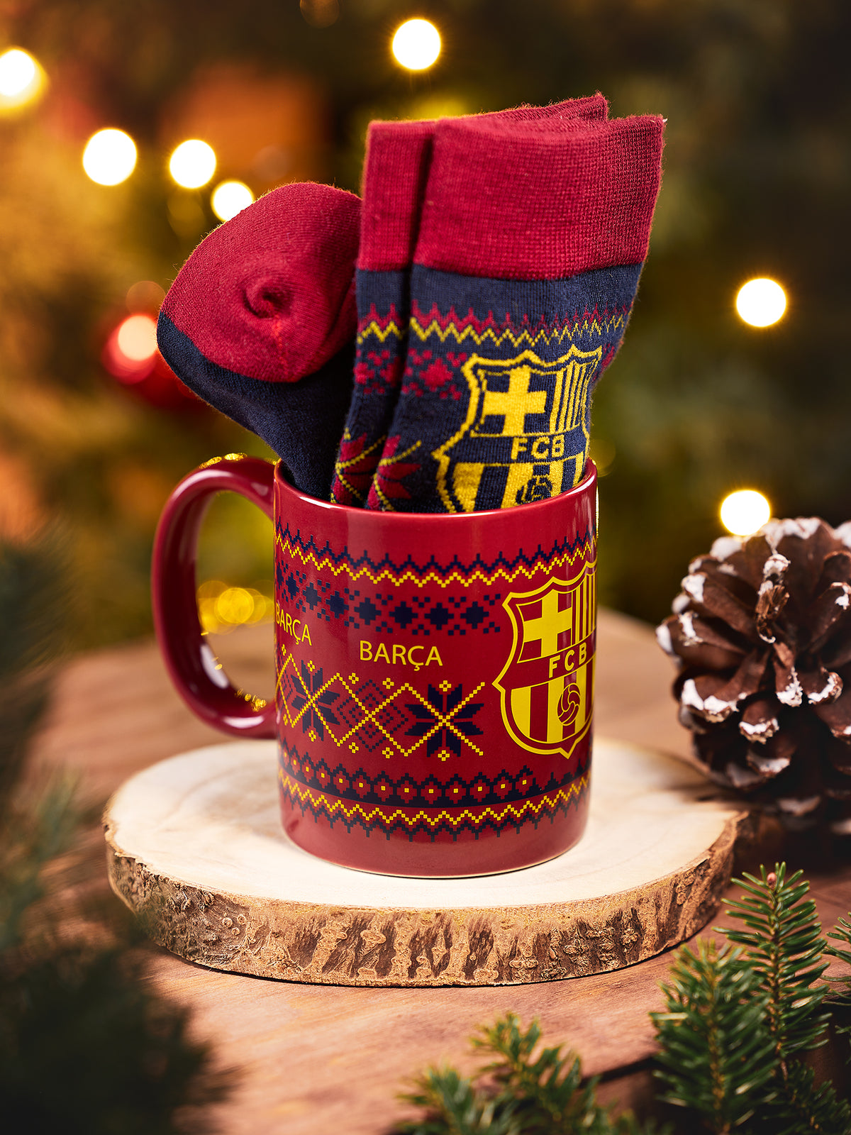 Taza roja del Barça con el escudo amarillo de FC Barcelona, sujetando calcetines plegados marino y rojo, sobre una rodaja de madera con luces festivas al fondo.