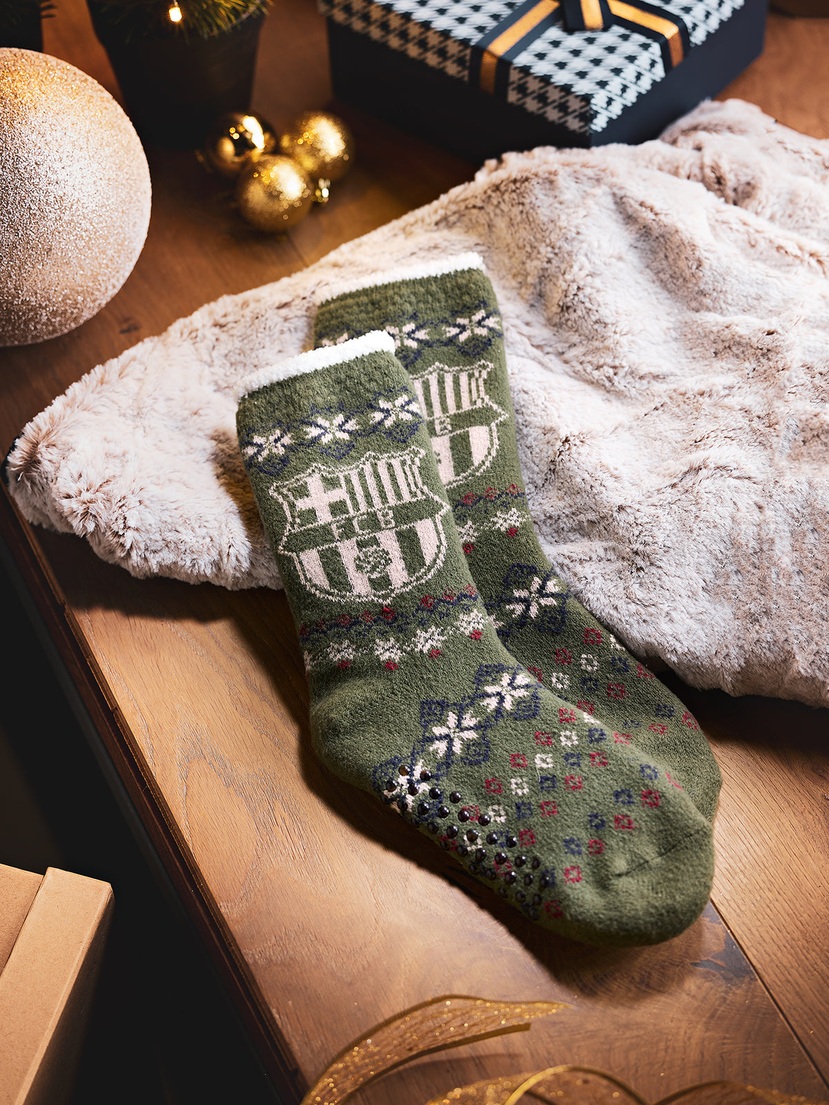 Paire de chaussettes vert olive FC Barcelona avec revers à motif et motifs d’écusson répétés, posées sur une couverture moelleuse avec des boîtes cadeaux et des ornements dorés.